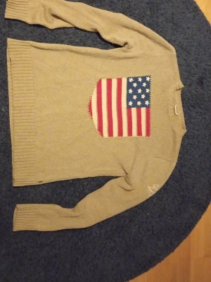 Beige stickad tröja Gant - Säljer en beige stickad tröja från Gant med stor amerikansk flagga på bröstet och broderad logga på ärmen. Tröjan har ribbade muddar och är långärmad. Perfekt för dig som gillar klassisk preppy stil.