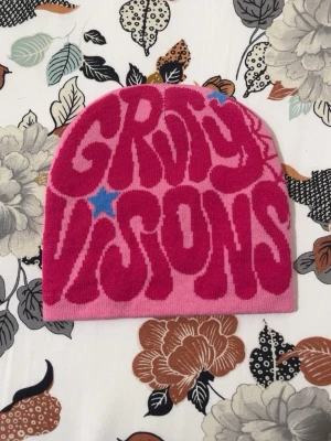 Rosa mössa med Groovy Visions-tryck - Säljer en rosa mössa med coolt psykedeliskt 'Groovy Visions'-tryck i mörkrosa och blått. Mössan har en klassisk form och är stickad i mjukt material, perfekt för dig som vill sticka ut med din stil.