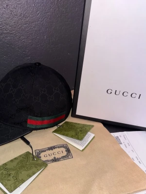 Svart Gucci keps med GG-mönster - Säljer en svart keps från Gucci med det klassiska GG-monogrammet över hela kepsen. Framtill finns en grön och röd rand som signaturdetalj. Kepsen är i tygmaterial och har justerbar rem bak. Perfekt för dig som gillar exklusiva accessoarer.