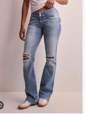 Ljusblå bootcut jeans med slitningar - Säljer ett par ljusblå bootcut jeans med slitningar över knäna och snygg tvätt. Jeansen har klassisk femficksdesign, hög midja och stängs med dragkedja och dubbla knappar. Perfekta för en avslappnad och trendig look.
