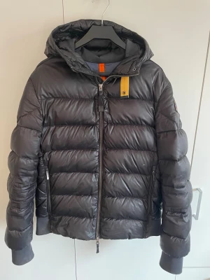 Svart jacka från Parajumpers - Snygg svart dunjacka från Parajumpers, modell Sheen. Jackan har huva, dragkedja framtill, två sidofickor med dragkedja och en gul detalj vid halsen. Perfekt för kalla vinterdagar och riktigt skön att bära. Litet hål finns kolla bild