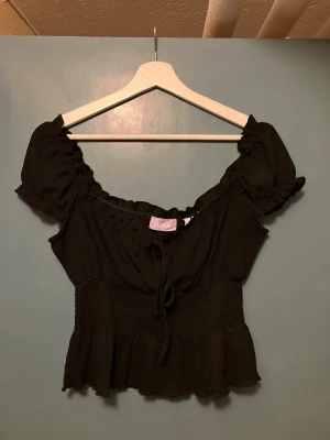 Svart smockad topp från JJXX - Trendig svart topp från JJXX med smockad midja, volangkant nedtill och korta puffärmar. Toppen har en söt knytning framtill och är tillverkad i ett mjukt, lätt material som passar perfekt till jeans eller kjol.