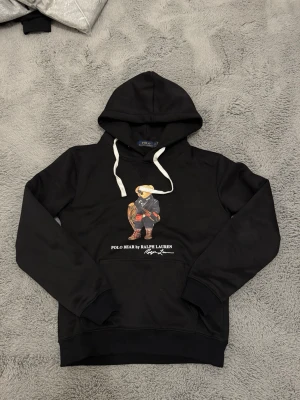 Svart hoodie Polo Bear Ralph Lauren - Svart hoodie från Ralph Lauren med den ikoniska Polo Bear-trycket framtill. Hoodien har vita dragsnören och en stor ficka på magen. Materialet känns mjukt och bekvämt, perfekt för en avslappnad stil. Snygg detalj med björnen i vinteroutfit och logga under.