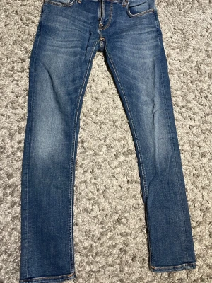 Blå jeans från Nudie Jeans - Säljer ett par klassiska blå  jeans från Nudie Jeans med snygga slitningar och kontrastsömmar. Jeansen har fem fickor och en smal passform. Perfekta för en trendig och avslappnad stil. Ganska små i storleken W32 L32 