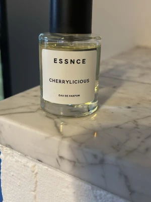 ESSNCE Cherrylicious Eau de Parfum - Fräsch parfym från ESSNCE med doften Cherrylicious. Kommer i en rund, genomskinlig glasflaska med svart lock och enkel vit etikett. Innehållet är ljusgult och flaskan har en modern, minimalistisk design.