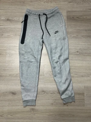 Gråa Nike byxor - Snygga ljusgrå mjukisbyxor från Nike med svart dragsko i midjan och en cool svart dragkedja längs ena fickan. Byxorna har ribbade muddar vid bensluten och Nike-logga på låret. Perfekta för chill eller träning.