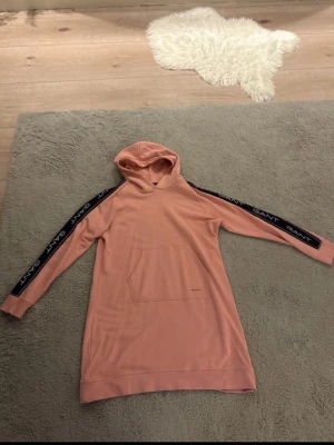 Rosa hoodieklänning från Gant - Säljer en rosa hoodieklänning från Gant med huva och känguruficka framtill. GANT-logga längs ärmarna. Mjuk. Perfekt för en avslappnad och trendig look.