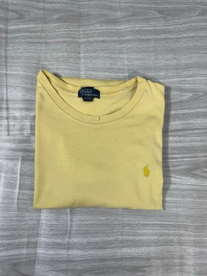 Gul t-shirt från Polo Ralph Lauren - Snygg gul t-shirt från Polo Ralph Lauren med klassisk rund hals och den ikoniska lilla broderade loggan på bröstet. Tillverkad i mjuk bomull för en skön känsla och enkel stil. Perfekt till jeans eller shorts för en clean look.