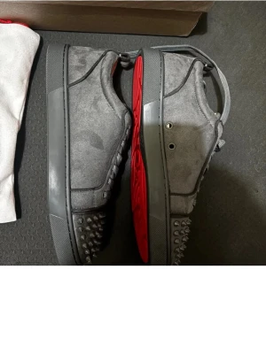 Gråa Louboutin sneakers med nitar - Säljer ett par gråa sneakers från Christian Louboutin i mocka med ikoniska röda sulor och coola nitar på tån. Skorna har lågt skaft, matchande grå skosnören och metalliska öljetter. Perfekta för dig som vill sticka ut med en edgy och lyxig stil.