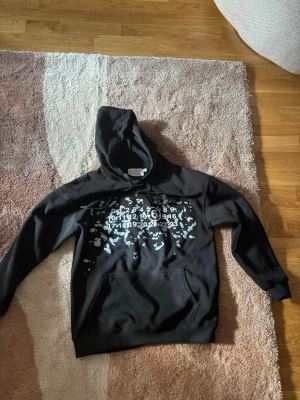 Svart hoodie Maison Margiela S - Svart hoodie från Maison Margiela med vit splatter-print och ikoniska siffror på bröstet. Klassisk känguruficka och justerbar huva med snören. Tillverkad i mjuk bomull för skön känsla och avslappnad passform.