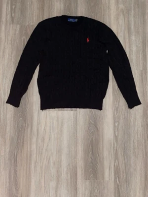 Svart kabelstickad tröja Polo Ralph Lauren - Kabelstickad svart tröja från Polo Ralph Lauren med röd broderad logga på bröstet. Tröjan har rund halsringning, långa ärmar och ribbade muddar vid ärmslut och nederkant. Klassisk och tidlös design i mjukt material.