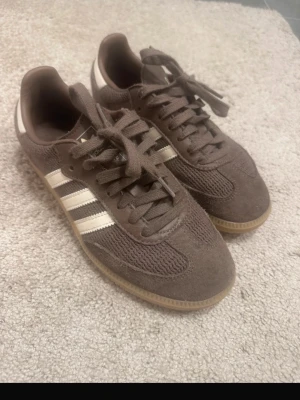 Bruna Adidas Samba sneakers - Säljer ett par bruna Adidas Samba sneakers med klassiska vita ränder på sidan. Skorna har ovandel i mocka och mesh, samt gummisula. Perfekt för dig som gillar retrostil och vill ha något som sticker ut lite extra. Köpta på Nelly 