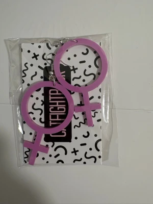 Lila Venus-symbol örhängen - Säljer ett par statementörhängen i form av Venus-symboler från Catfightback. De är lila och gjorda i lätt plastmaterial med silvriga krokar. Perfekta för dig som vill göra ett starkt intryck och visa din stil.
