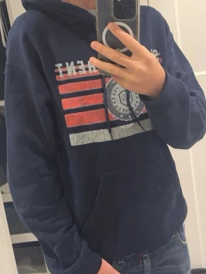 Saint Laurent hoodie - Mörkblå hoodie med huva och känguruficka. Framsidan har ett stort tryck i vitt, rött och grått med text och grafiskt mönster. Hoodien har dragsko i huvan och ribbade muddar vid ärmslut och nederkant. Perfekt för en avslappnad och cool stil.