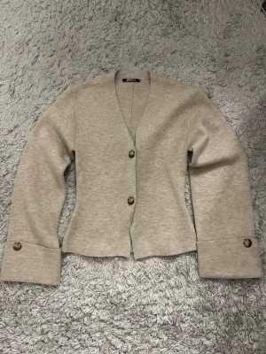 Beige kofta från Gina Tricot - Stilren beige kofta från Gina Tricot med v-ringning och två stora knappar framtill. Koftan har långa ärmar med breda muddar och knappdetaljer vid ärmsluten. Perfekt att slänga över en t-shirt för en chill look.