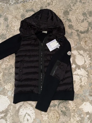 Svart dunväst med huva från Moncler - Snygg svart dunväst från Moncler med huva och stickade svarta ärmar. Västen har dragkedja framtill, två sidofickor med dragkedja och en Moncler-logga på ärmen. Materialet är en mix av dun och stickat tyg för en modern look. Helt ny jacka för året om. Skriv gärna över den minsta lilla funderingen.