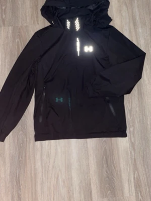 Svart vindjacka från Under Armour - Snygg svart vindjacka från Under Armour med huva och reflekterande detaljer på bröstet. Jackan har dragkedja framtill, elastiska muddar och två sidofickor. Perfekt för blåsiga dagar och sportiga outfits.