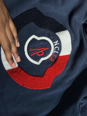 Mörkblå sweatshirt från Moncler  - Snygg mörkblå sweatshirt från Moncler med stort broderat logomärke i rött, vitt och blått på bröstet. Tröjan har rund halsringning och ribbade muddar. Perfekt för dig som gillar streetwear och exklusiva märken. Den kan sita lite på kropen men är parfekt för dig som gilar mer slim tröjor