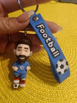 Nyckelring med fotbollsspelare blå - Säljer en söt nyckelring med en liten figur i blå fotbollströja och nummer 10, samt en blå rem med texten 'Football' och en fotboll. Perfekt accessoar för dig som älskar fotboll! Materialet är mjuk plast/gummi. Helt ny. Oanvänd 