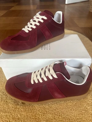 Vinröd Maison Margiela Gats - Snygga bordeauxfärgade sneakers från Maison Margiela med ovandel i mocka och läder, vita snören och klassisk gummisula. Ikonisk design med låg profil och vita detaljer på insidan. Perfekta för dig som vill ha något stilrent men ändå unikt.