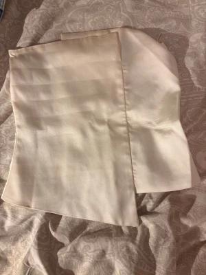 Champagnefärgad satin korsettopp - Snygg axelbandslös korsettopp i champagnefärgad satin med asymmetrisk nederkant och dragkedja bak. Perfekt för dig som vill ha en trendig och elegant look. Materialet är glansigt och ger en lyxig känsla.