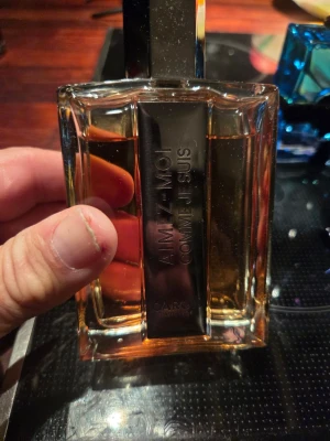 AIMEZ-MOI Comme Je Suis parfym - AIMEZ-MOI Comme Je Suis från Caron är en exklusiv parfym i en elegant, rektangulär glasflaska med metallisk detalj framtill. Flaskan har en varm bärnstensfärgad vätska och en modern, stilren design som känns lyxig och tidlös. 90 % ej box