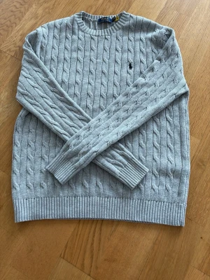 Ljusgrå kabelstickad tröja Polo Ralph Lauren - Snygg ljusgrå kabelstickad tröja från Polo Ralph Lauren med rund halsringning och diskret broderad logga på bröstet. Tröjan har långa ärmar och ribbade muddar vid ärmslut och nederkant. Perfekt för lager på lager och klassisk stil. Knappt använd as snygg nu till våren. 