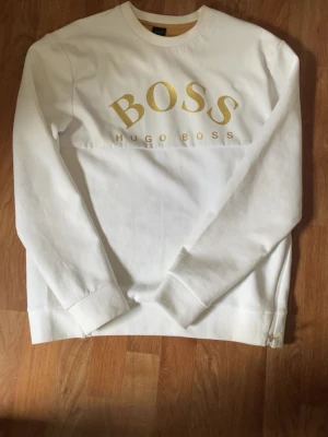 Vit Hugo Boss hoodie med gulddetaljer - Snygg vit hoodie från Hugo Boss. Nytt skick då jag säljer den väldigt billigt. Ny pris 999kr. Pris kan diskuteras.