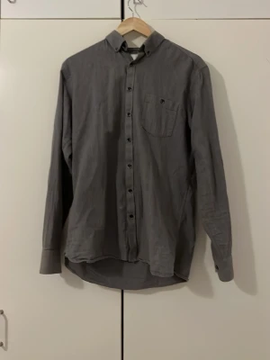 Mörkgrå skjorta med knappdetaljer (M) - Mjuk och härlig grå skjorta med klassisk krage och bröstficka. Skjortan har långa ärmar och knappar hela vägen framtill. Perfekt för dig som gillar en enkel men snygg look. Tillverkad i mjuk bomull för skön känsla. Du kan ha den under en lång rock, kavaj eller helt enkelt till vardags. Om du handlar hos mig kan vi alltid diskutera priset! 