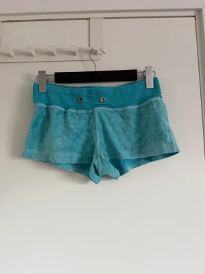 Turkosa frottéshorts med ribbad midja - Säljer ett par turkosa shorts i mjuk frotté med bred ribbad midja och två dekorativa metallöljetter framtill. Perfekta för sommardagar eller som chill-plagg. De har en relaxed passform och är riktigt bekväma. Juicy coutur liknande