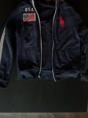 Marinblå hoodie från Ralph Lauren - Snygg marinblå hoodie från Ralph Lauren med dragkedja, vita detaljer och två fickor framtill. På bröstet finns ett stort rött Polo-logotryck, USA-text och en broderad amerikansk flagga. Perfekt för dig som gillar sportig och klassisk stil.