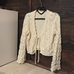 Beige stickad kofta från Jolio&Co - Säljer en beige stickad kofta från Jolio&Co / köpt på Missäy, med fina virkade detaljer och knytband i midjan. Koftan har lång ärm, vågig kant och ett romantiskt mönster som ger en söt vintagekänsla. Andvänd en gång. Köpte för 499.