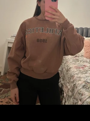 Brun sweatshirt med tryck 'ATHLETICS 1998' - Cool brun sweatshirt med texten 'ATHLETICS 1998' i vitt och svart tryck på bröstet. Oversized passform med ribbade muddar vid ärmslut och nederkant. Perfekt för en avslappnad och sportig look.