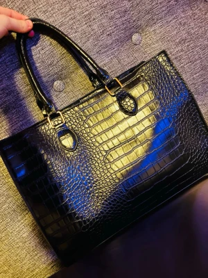 Svart handväska i krokodilimitation - Snygg svart handväska i glansigt konstläder med krokodilpräglat mönster. Väskan har två stadiga handtag med guldfärgade detaljer och rymligt innerfack med dragkedja. Perfekt för dig som gillar trendiga accessoarer med lyxig känsla.