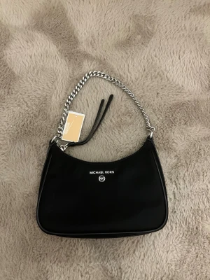 Svart Michael Kors handväska - Stilren svart handväska från Michael Kors med silverfärgad kedjerem och logga framtill. Väskan är i slät konstläder och har en enkel, avlång form. Perfekt för dig som gillar en klassisk och trendig look.