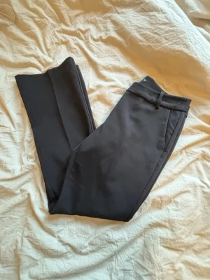 Svarta kostymbyxor som är lite utsvängda  - Snygga svarta kostymbyxor som är lite utsvängda och är i short. Byxorna har hällor för skärp och sidofickor. Perfekta för en stilren look.  Materialet känns slätt och dressat. 