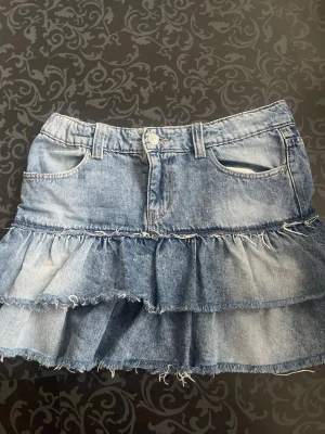 Blå volangkjol i denim - Säljer en blå jeanskjol med två volanglager och råa kanter för en cool, avslappnad look. Kjolen har klassiska fickor fram och bak samt bälteshällor. Perfekt för dig som gillar denim och vill ha en trendig twist på din outfit.