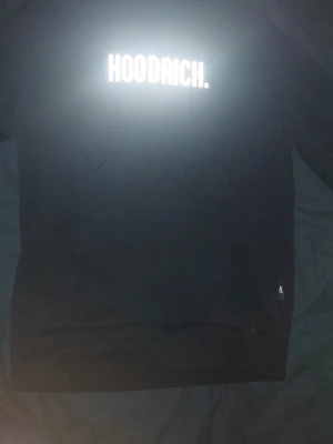 Svart hoodie från Hoodrich - Snygg svart hoodie från Hoodrich med stor vit logga på bröstet. Hoodien har en klassisk huva med dragsko och en stor ficka framtill. Tillverkad i mjukt material som passar perfekt för en avslappnad streetwear-look.