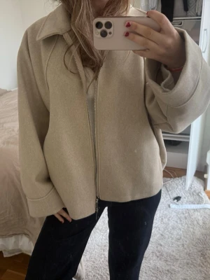 Beige ulljacka med dragkedja - Snygg beige jacka i ullmix med dragkedja framtill och bred krage. Jackan har en loose passform och långa ärmar, perfekt för lager på lager. Enkel och stilren design som passar till det mesta.