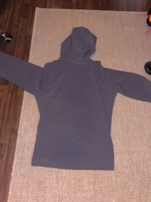 Mörkgrå vindjacka från Under Armour - Snygg mörkgrå vindjacka från Under Armour med huva och dragkedja framtill. Jackan har två sidofickor och en diskret logga på bröstet. Tillverkad i ett lätt och smidigt material som passar perfekt för blåsiga dagar.