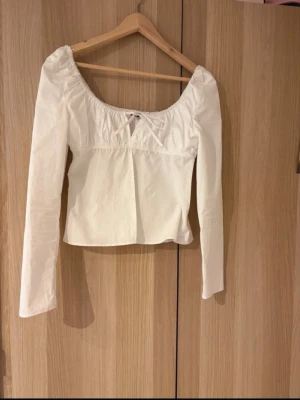 Vit långärmad topp med rosett - Söt vit topp med lång ärm och rundad urringning. Toppen har en liten rosett framtill och dragkedja i sidan för enkel påklädning. Passformen är något croppad och materialet känns som bomull. Perfekt för en trendig och stilren look.