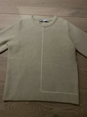 Beige stickad tröja från Zara - Stilren beige stickad tröja från Zara med rund halsringning och dekorativ vit söm framtill. Tröjan har långa ärmar och ribbade muddar vid ärmslut och nederkant. Perfekt för en enkel och trendig look.