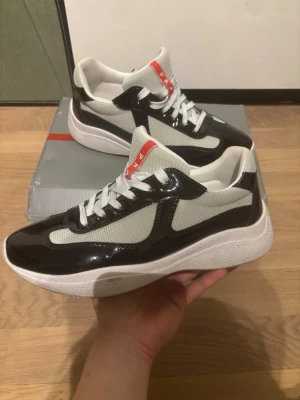 Prada sneakers svart och vit - Snygga Prada sneakers i svart lack och vit mesh med vita skosnören och röd logga på plösen. Tjock vit sula ger en modern vibe och kontrasten mellan materialen gör dem riktigt stilrena. Perfekta för dig som vill ha något exklusivt och trendigt. Box medföljer och rengörs så gott det går innan frakt 