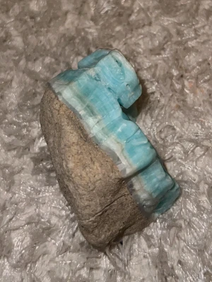 Rå larimar sten - Unik rå larimar sten i vackra blå och vita nyanser. Stenen har en naturlig, oregelbunden form och är delvis omgiven av beige berggrund. Perfekt för dig som gillar kristaller eller vill skapa egna smycken.