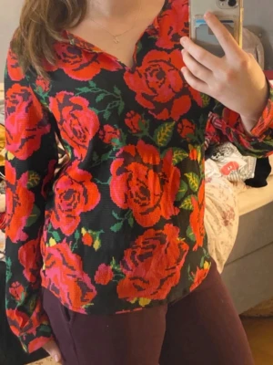 Blommig långärmad blus med rosor - Snygg blus med svart botten och stora röda och rosa rosor samt gröna blad. Blusen har V-ringning och långa ärmar. Materialet ser ut att vara mjukt och stretchigt, perfekt för dig som gillar färgstarka plagg med statement-mönster. Bra passform, normal i storleken och skönt material. 