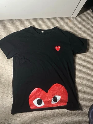 Svart Comme des Garçons Play t-shirt - Svart t-shirt från Comme des Garçons Play med ikoniskt rött hjärta med ögon tryckt både på bröstet och i större format nedtill. Klassisk passform och rund halsringning. Perfekt för dig som gillar streetwear och vill sticka ut med en unik detalj. Perfekt nu för sommaren. Obs: tröjan kommer endast.