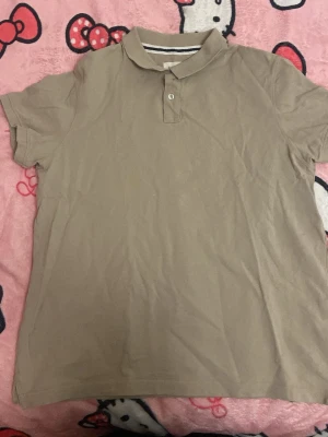 Beige pikétröja med krage - Klassisk beige pikétröja med korta ärmar och krage. Tröjan har två knappar vid halsen och en diskret randig detalj på insidan av kragen. Perfekt till en avslappnad och stilren look.