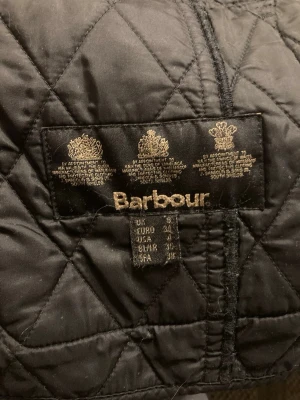 Svart quiltad dam jacka från Barbour - Svart damjacka från Barbour med knappar och guldig design runt fickorna och märket. Jackan har 4 fickor och det finns även hällor runt omkring jackan vid behov av snöre/bälte( Frivilligt). Jackan är knappt använd och är i perfekt skick.