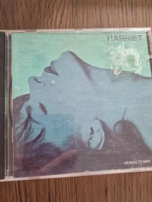 CD Harriet - Woman to Man - CD-skiva med albumet 'Woman to Man' av Harriet. Omslaget har en blågrön ton med ett konstnärligt porträtt i profil. Baksidan listar låtarna och har stilren textdesign. Perfekt för dig som gillar pop och samlar på fysiska album.