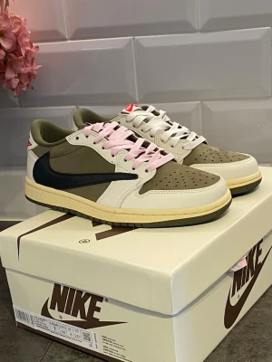 Nike Air Jordan 1 Low Travis Scott - Nike Air Jordan 1 Low Travis Scott med olivgröna, vita och beige paneler, svart omvänd Swoosh och rosa samt vita skosnören. Yttermaterial i läder och mocka, med röd logga på hälen och klassisk AJ1-sula. Snygg och unik sneaker för dig som gillar streetwear.
